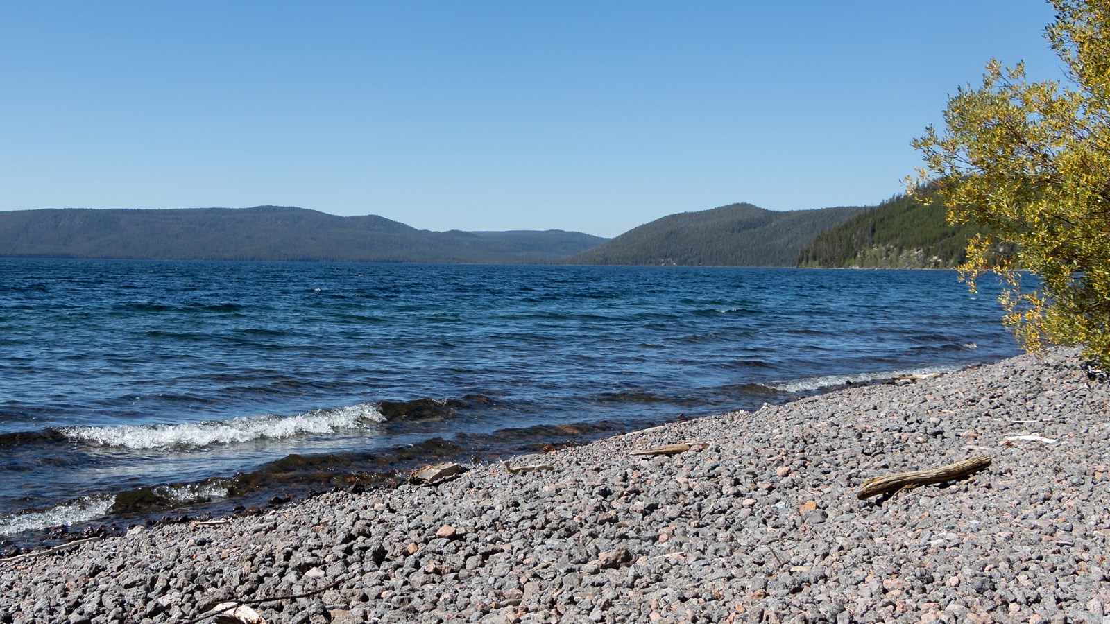 A rocky beach on a large, blue lake with forested mountain slopes in the distance.