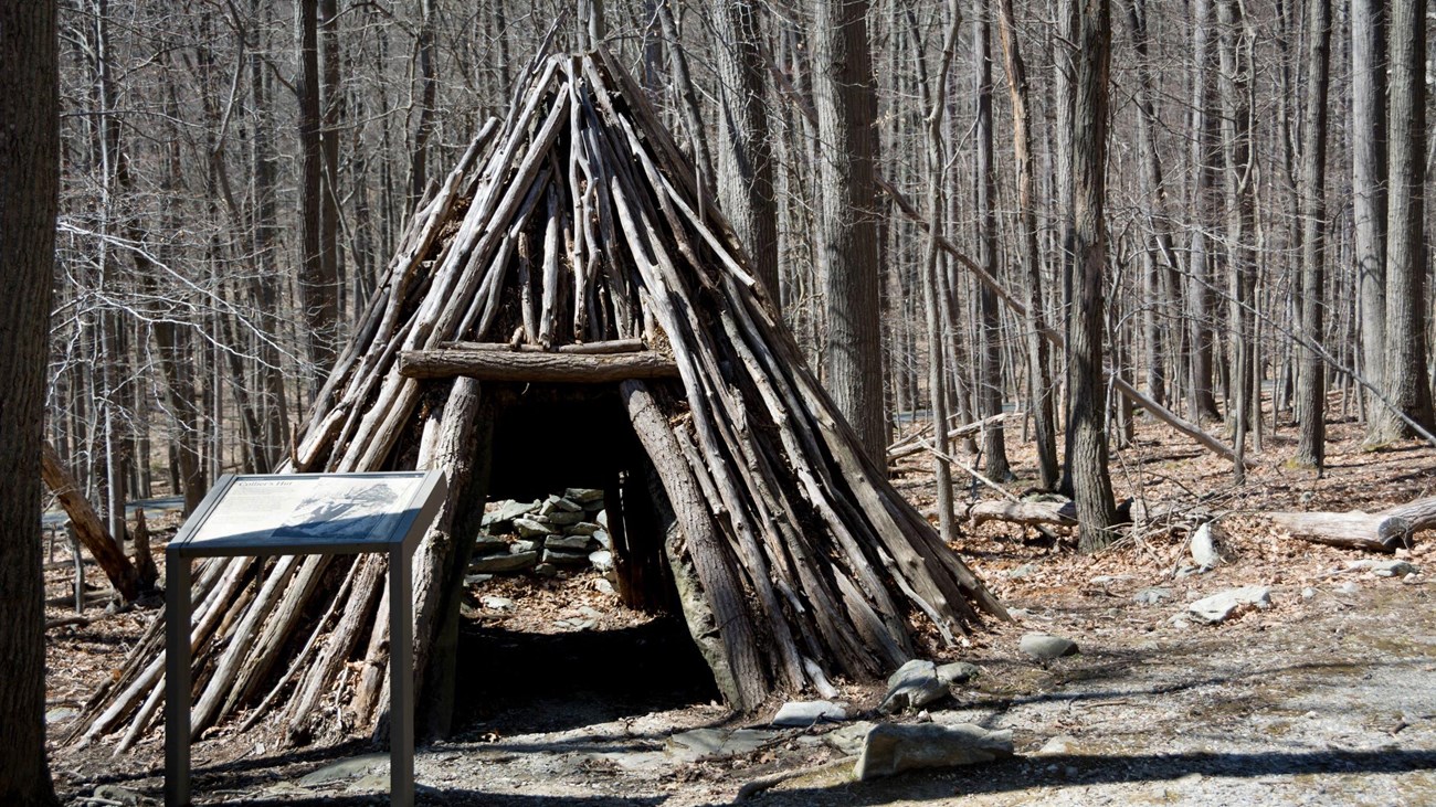 A triangular wooden hut made of sticks in a forest.
