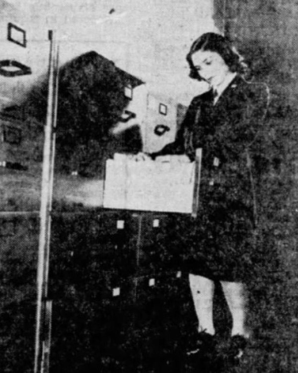 Grainy photo of woman in a dark skirt suit going through files at a wall of file cabinets.