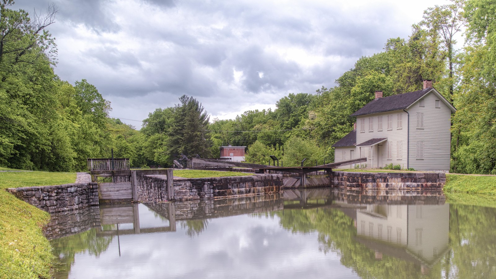 Water moves toward a white lockhouse and wooden lock. Trees line the side and background. 