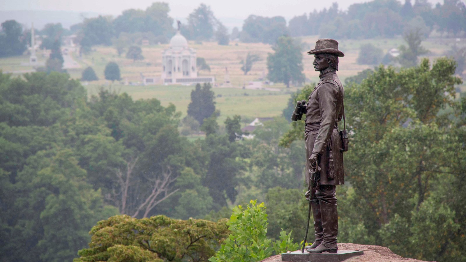 A bronze statue of a soldier on a rock with monuments, tour roads and trees behind him.