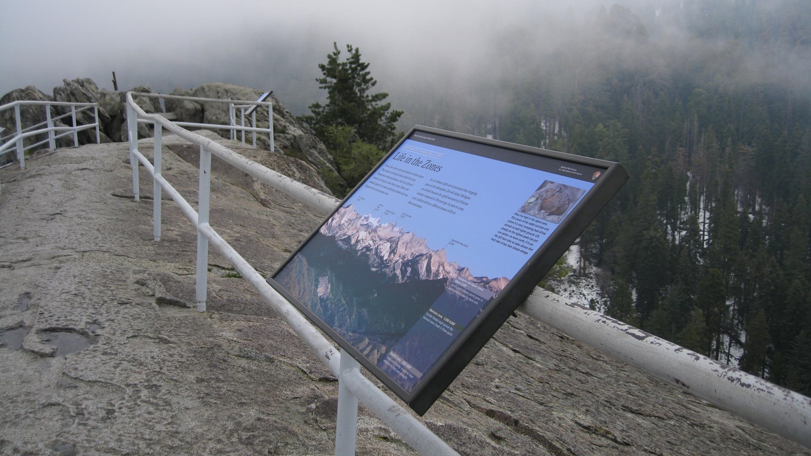 An exhibit sign is mounted to a white metal rail lining the steep trail on Moro Rock\'s peak.
