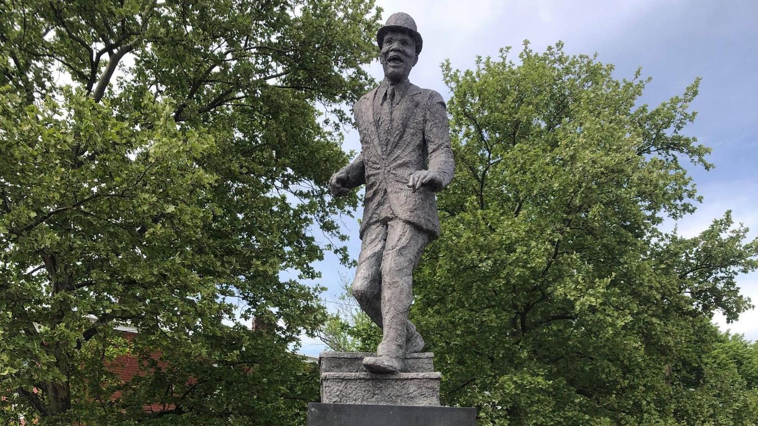 A metal statue of Bill Robinson mid-dance step, wearing a suit and bowler hat standing on a pillar