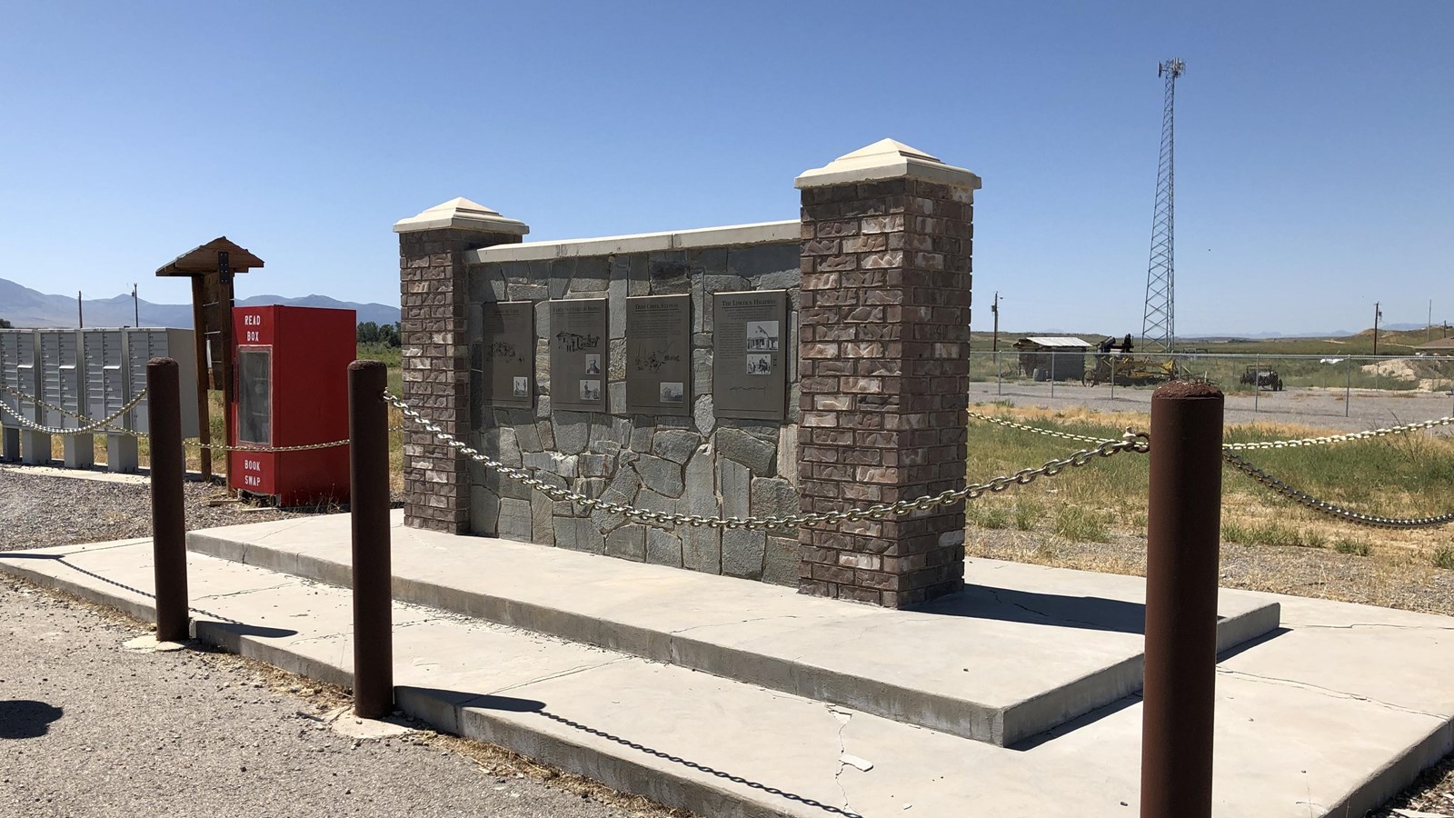 A chain barrier surrounds stone wall monument with 4 interpretive panels and brick posts on sides.