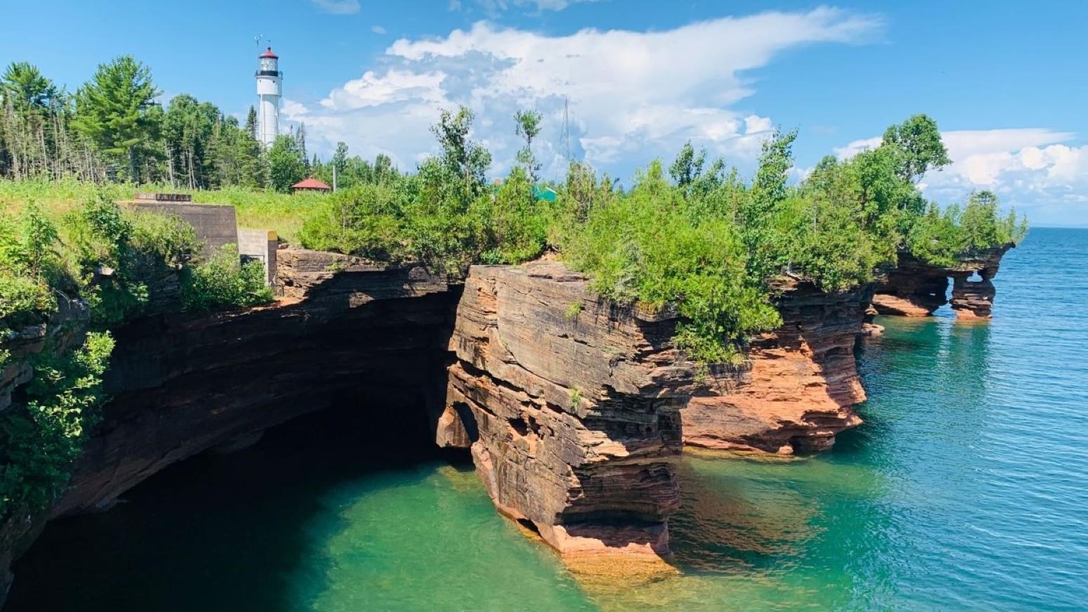 Brown sandstone cliffs covered with trees protrude out into the water with a lighthouse on top. 