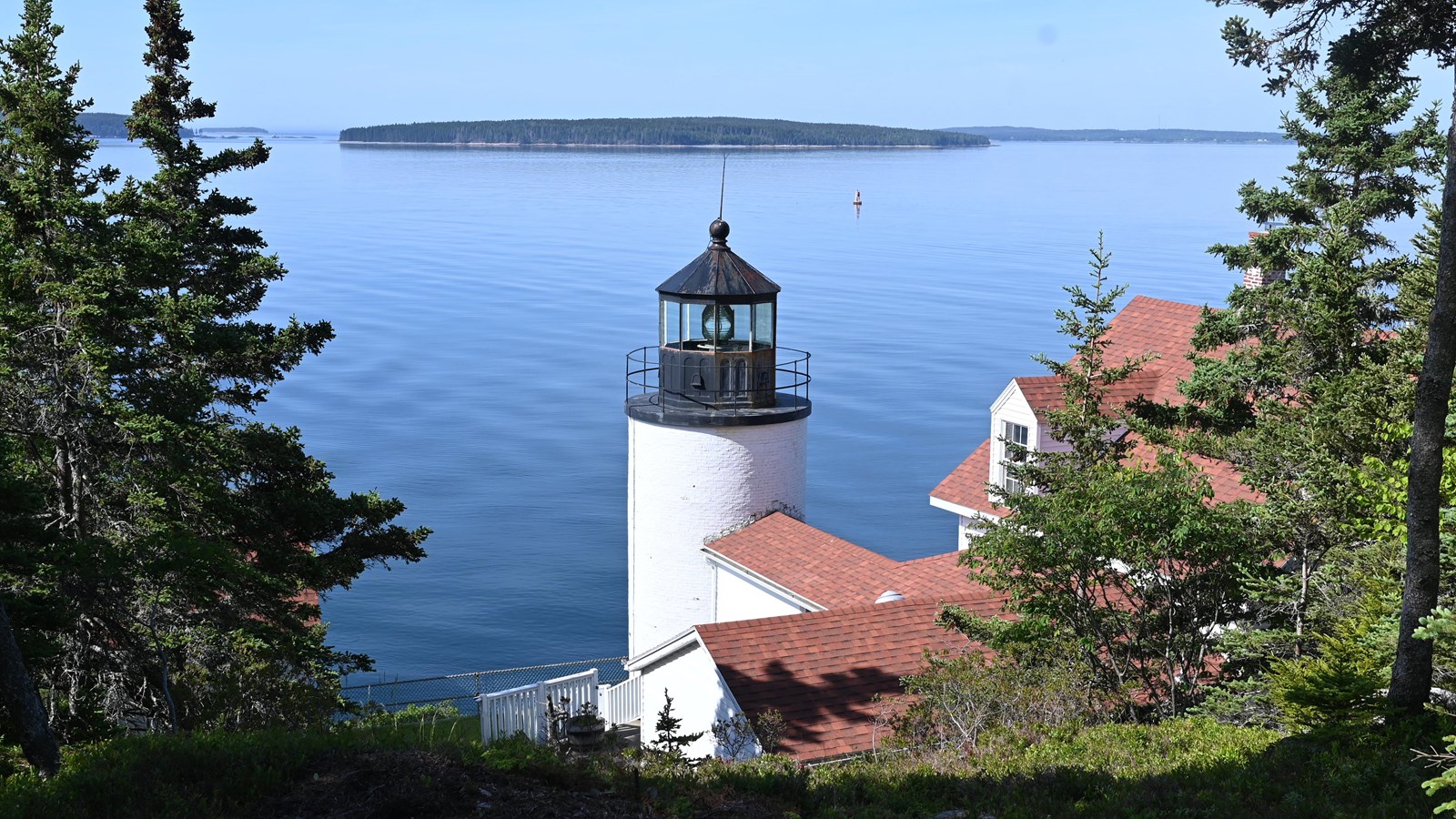 A white lighthouse with a black roof being viewed from above with the sea and an island behind it. 