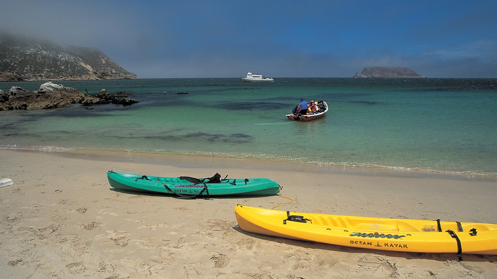 Kayaks on white sand beach with blue water and skiff with passengers approaching beach.