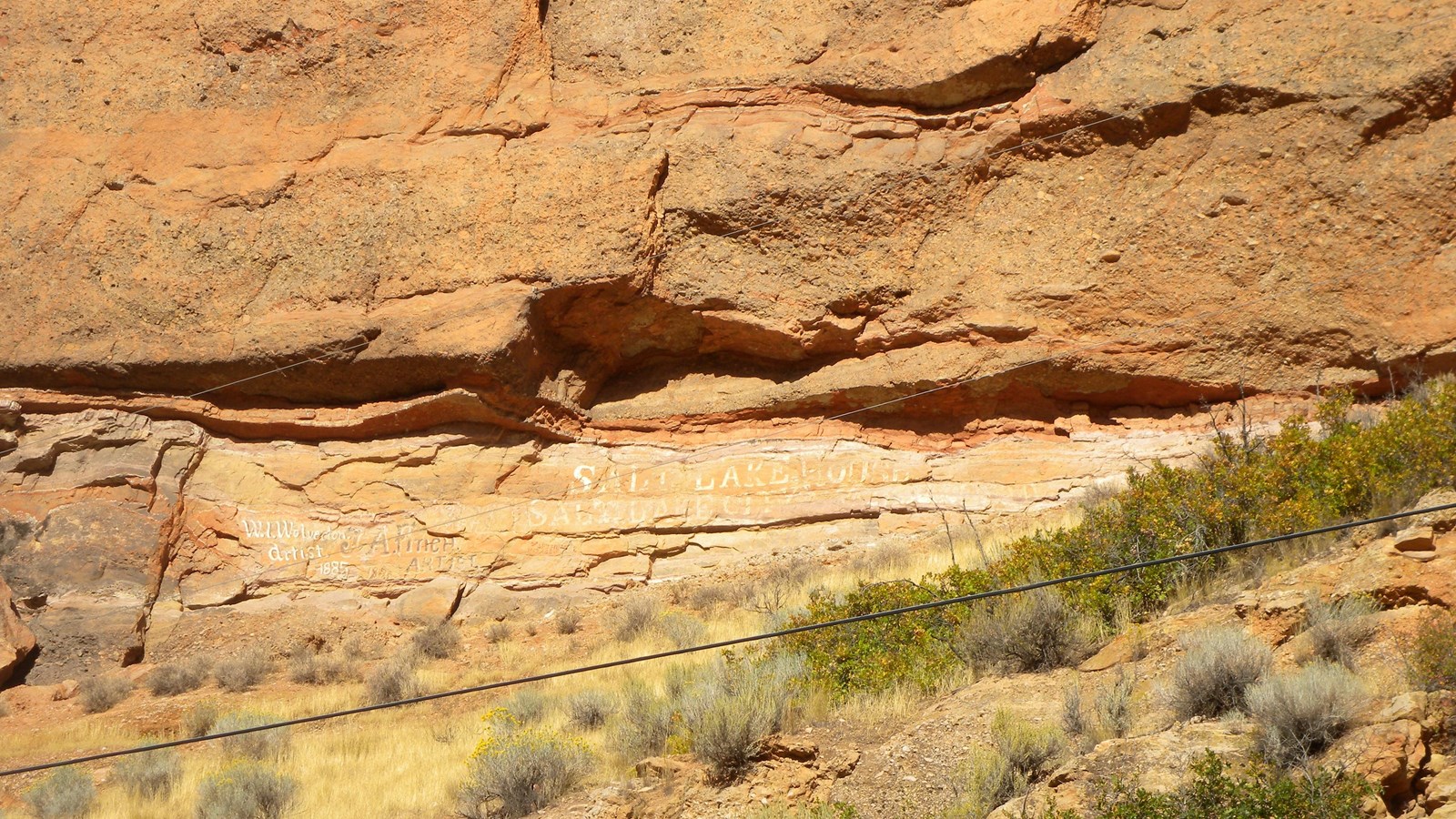 Painted letters run along the base of a rugged, red sandstone cliff face. 