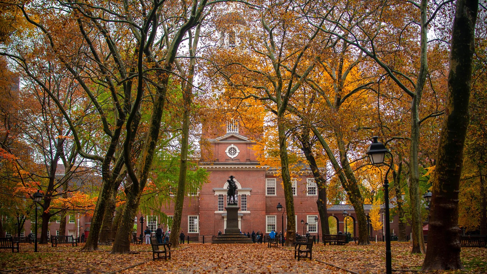 A color photo of a path surrounded by trees and fall foliage, Independence Hall is in the background