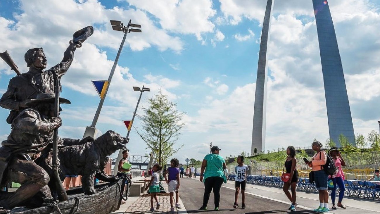  a group of people standing along a trail next to the Captain\'s Return statue in front of the Arch