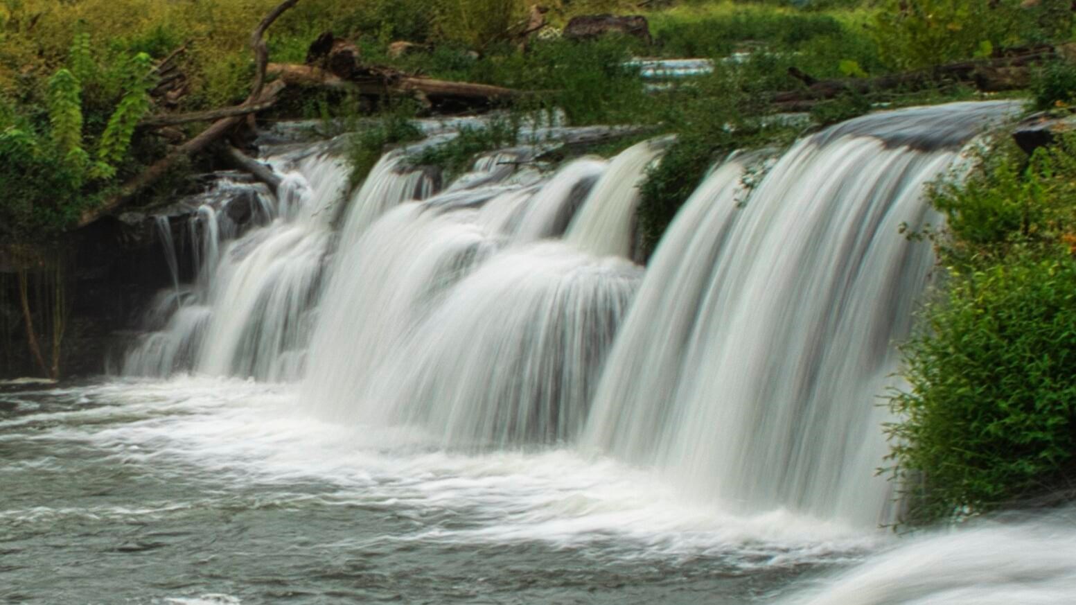 A wide river of water flowing 10 feet over rocks into a deep pool surrounded by vegetation and trees