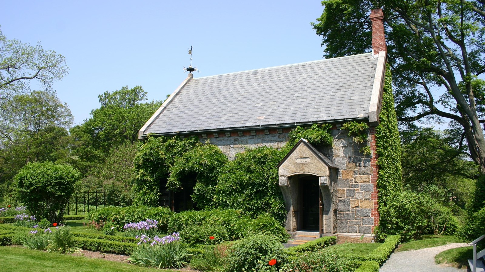 A stone and brick building with a chimney and weathervane. Wisteria vines climb up the sides.