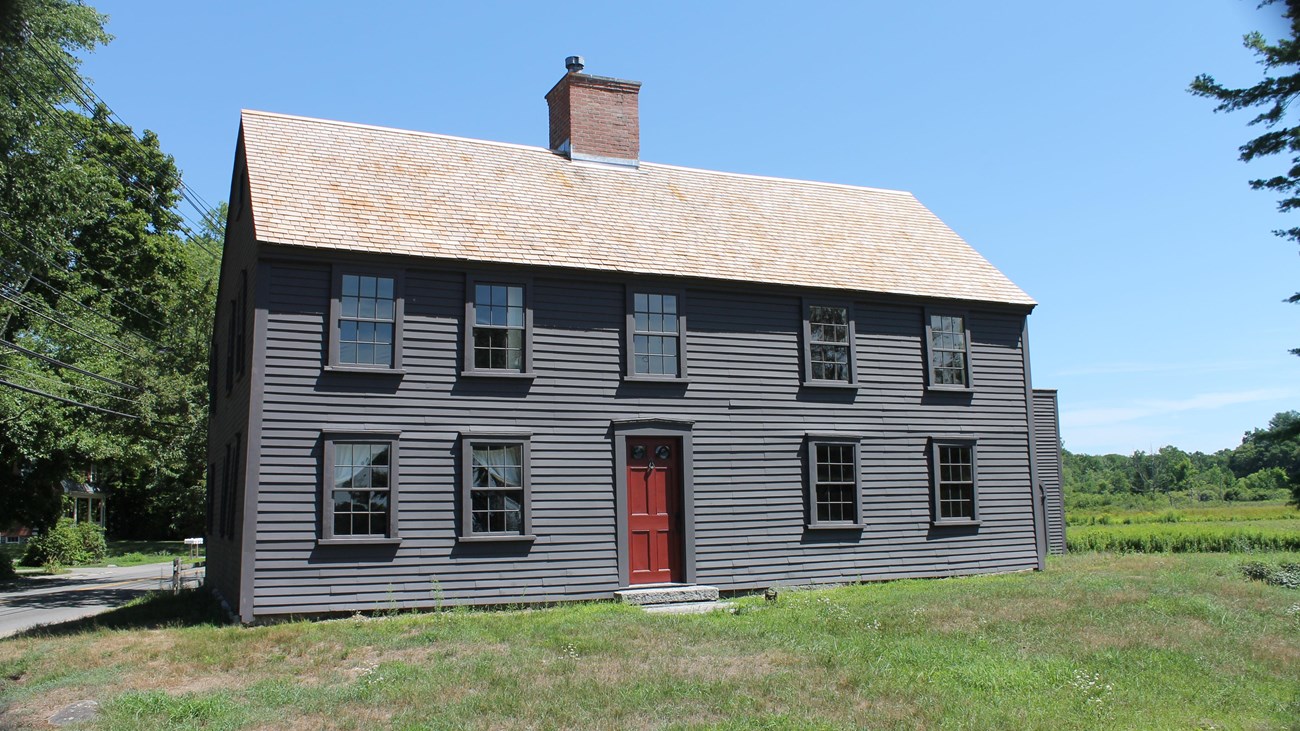 A two story salt box style home made of wood with a central chimney sits in an open grassy field