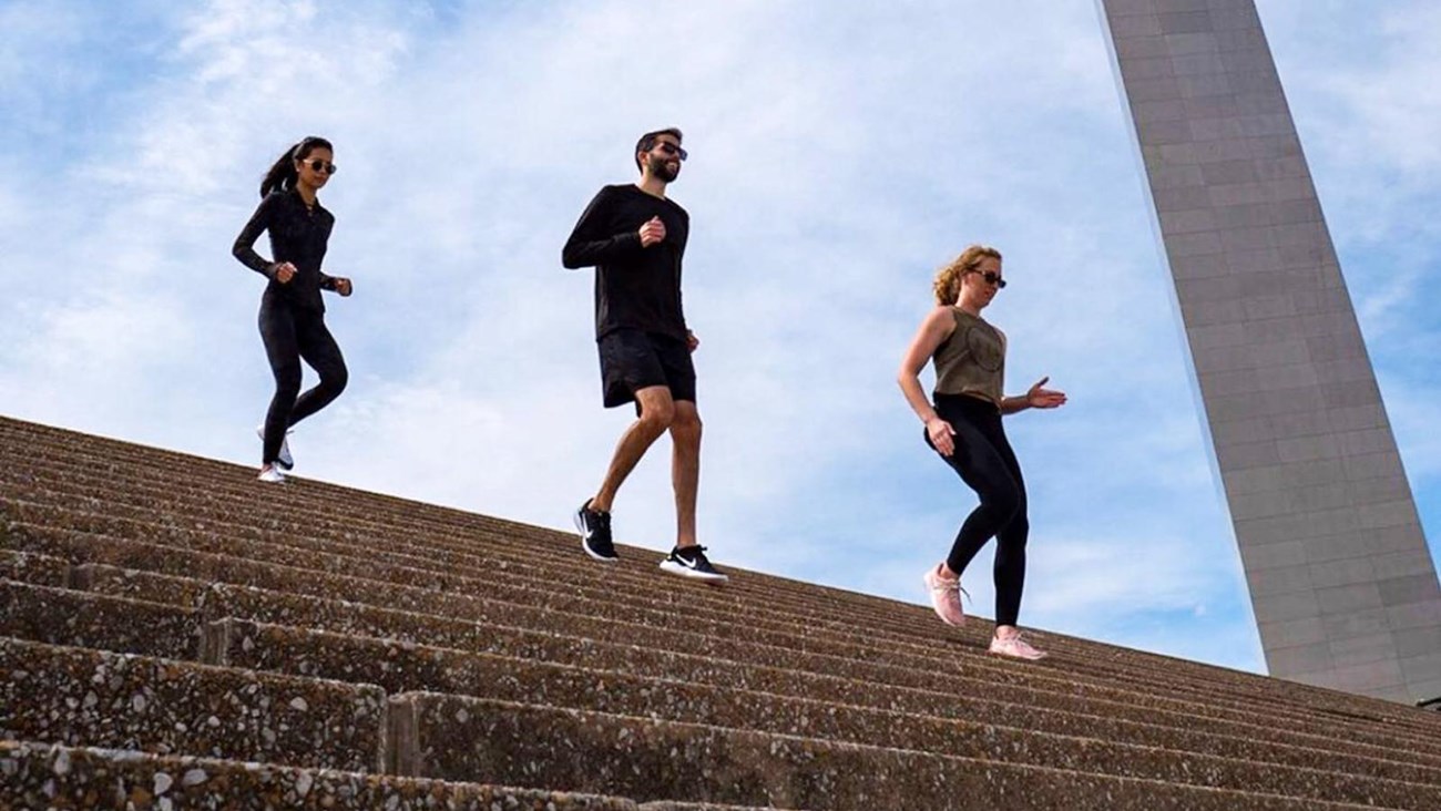 two women and a man jogging down the Grand Staircase in front of the Arch