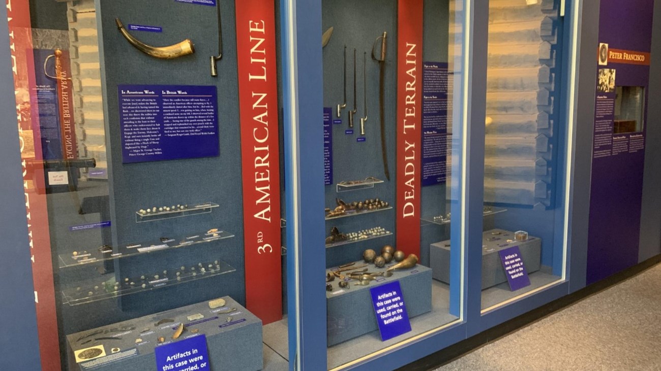 A museum exhibit with blue and red planning featuring weapons like musket balls & bayonets