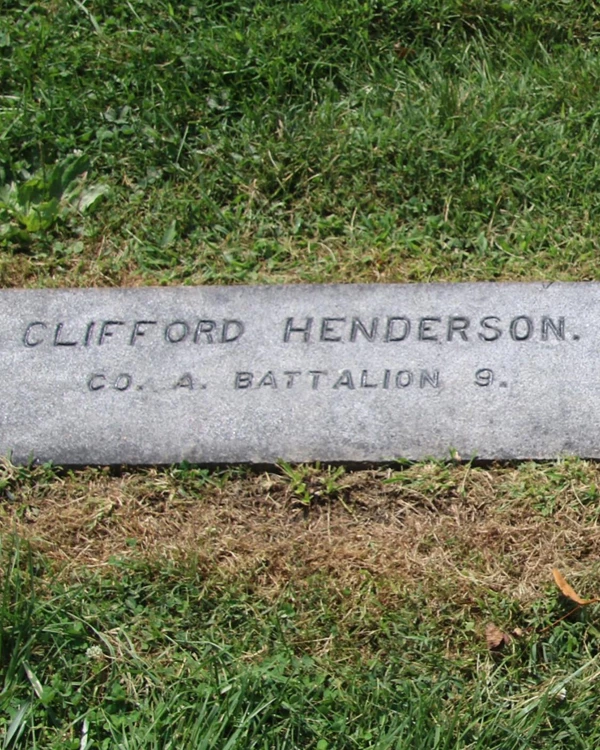 A dark colored gravestone engraved with the name Clifford Henderson lays flat among grasses.