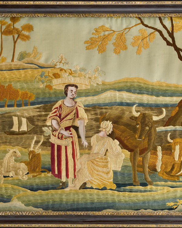Framed needlework piece of a kneeling woman milking a cow, another woman stands nearby. 