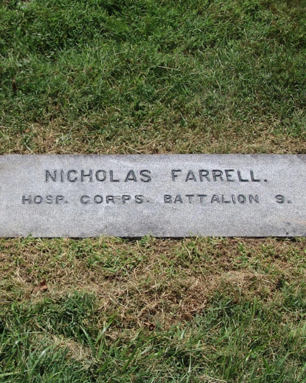 A dark gravestone is carved with the name of Nicholas Farrell lays flat on the ground among grass.