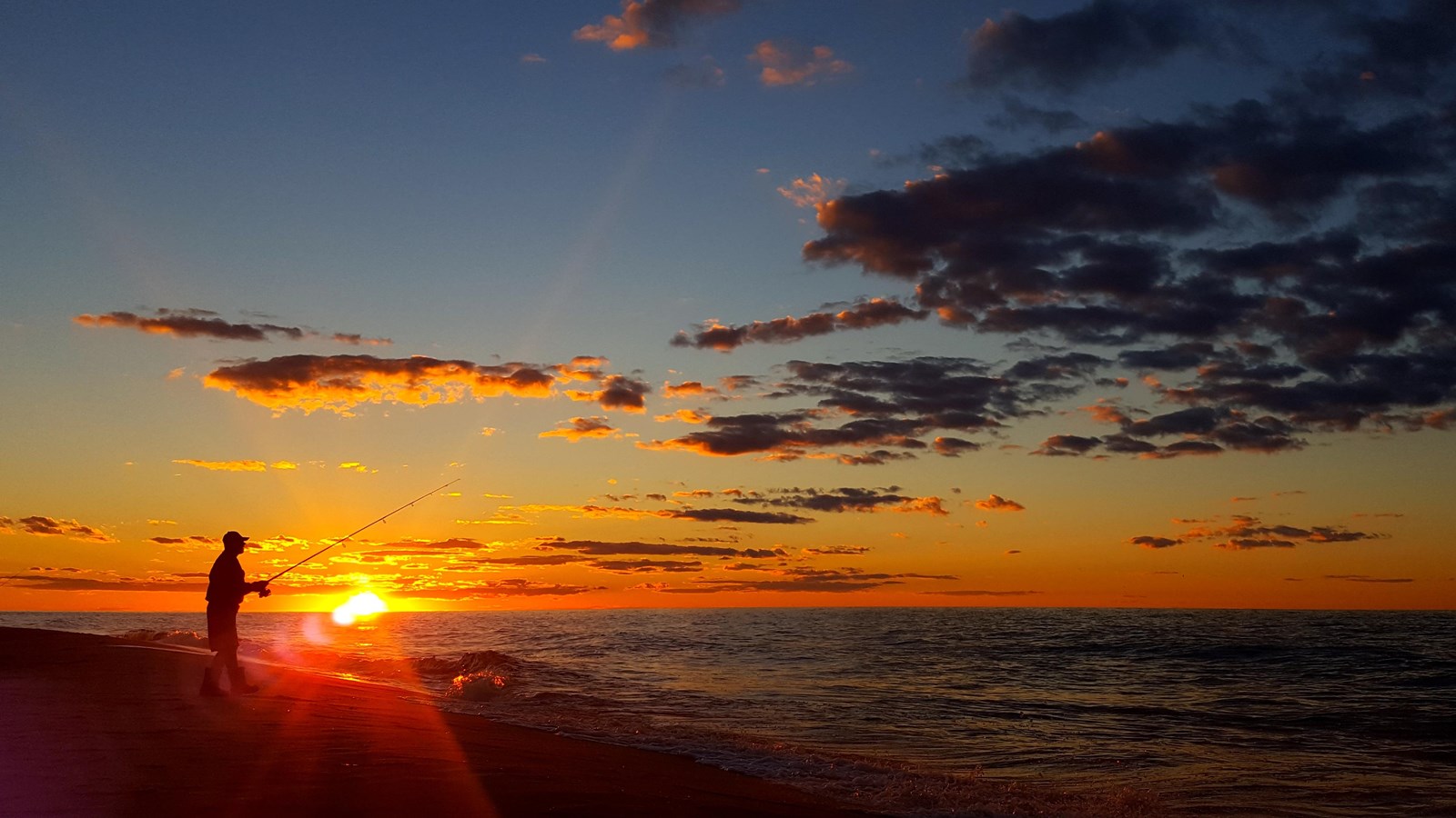 Person with a fishing pole silhouetted against a deep orange sky as the sun sets into the water.