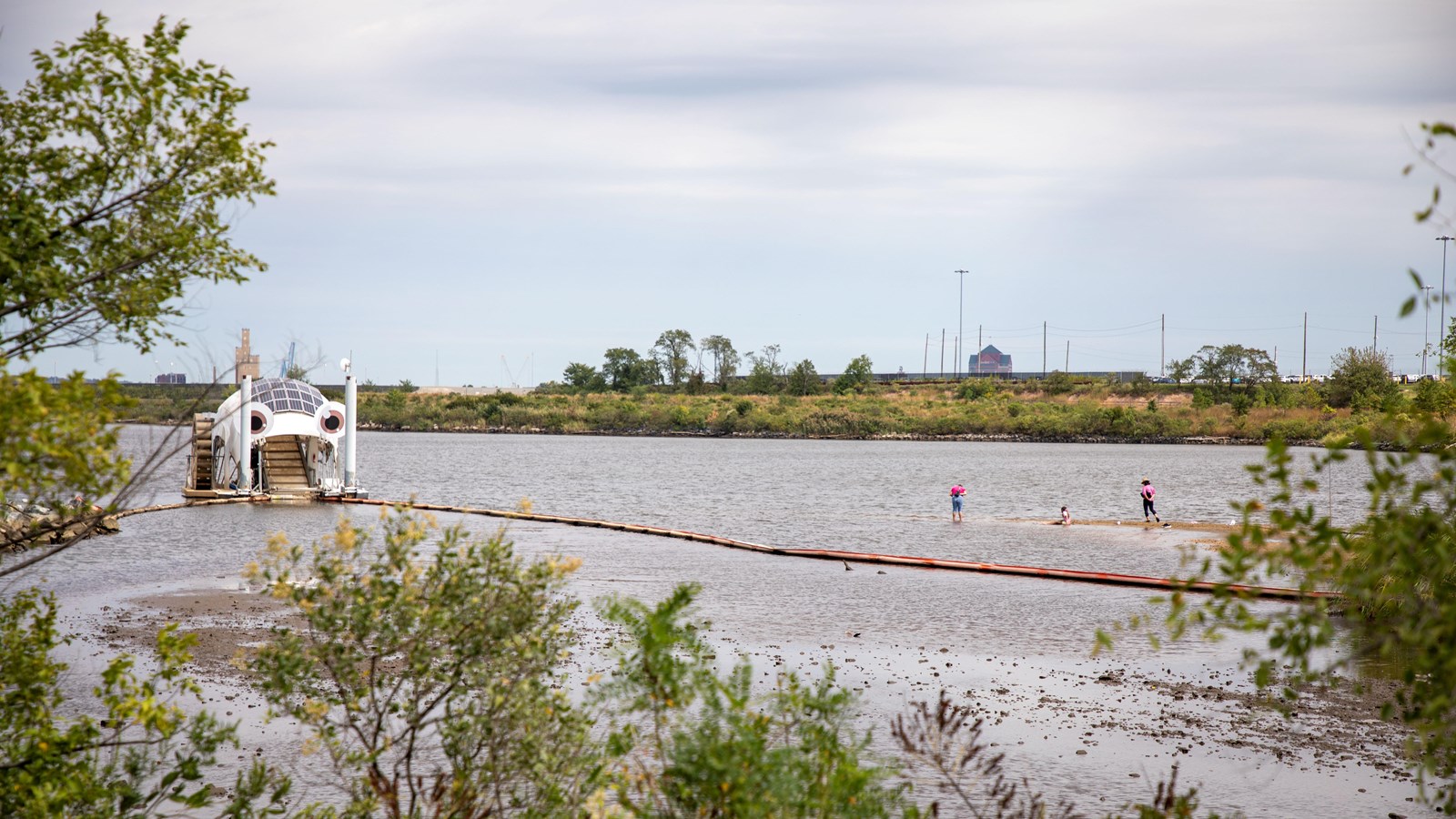 A white, dome-shaped structure in a river with people on a nearby sandbank surrounded by greenery.