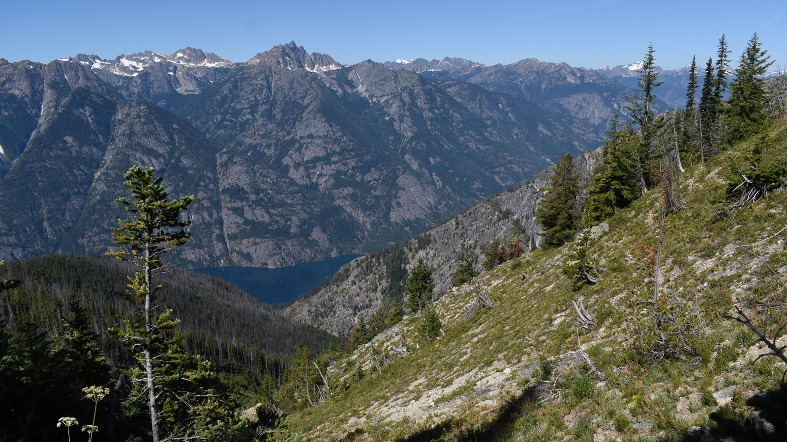 A view looking down past forested hills to steep mountains rising from a long, thin lake.