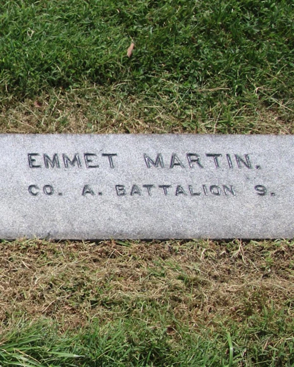 The dark colored gravestone engraved with the name Emmert Martin lays flat with the ground.