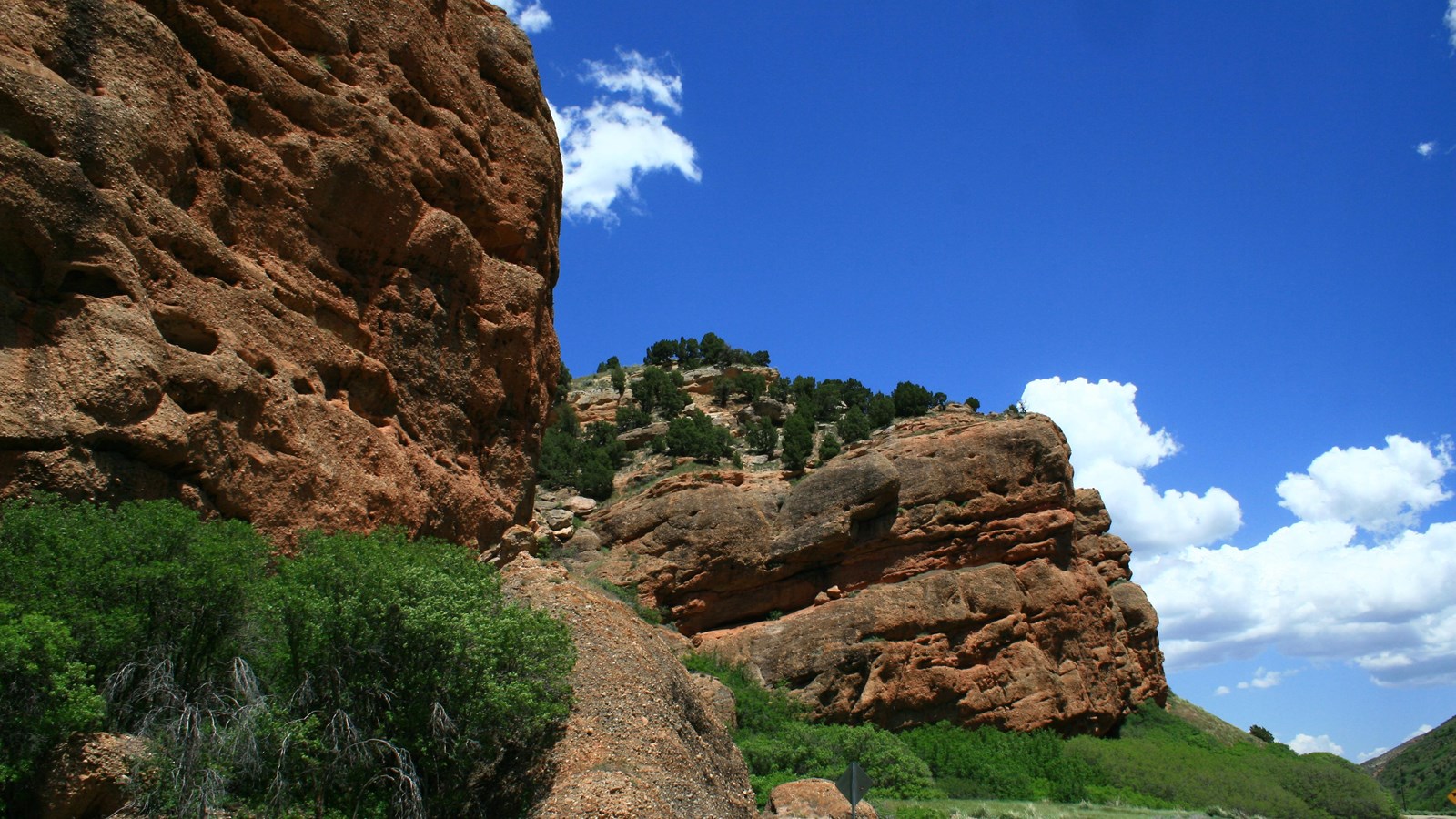 A large, red-hued rock outcropping hangs near a road through a canyon.