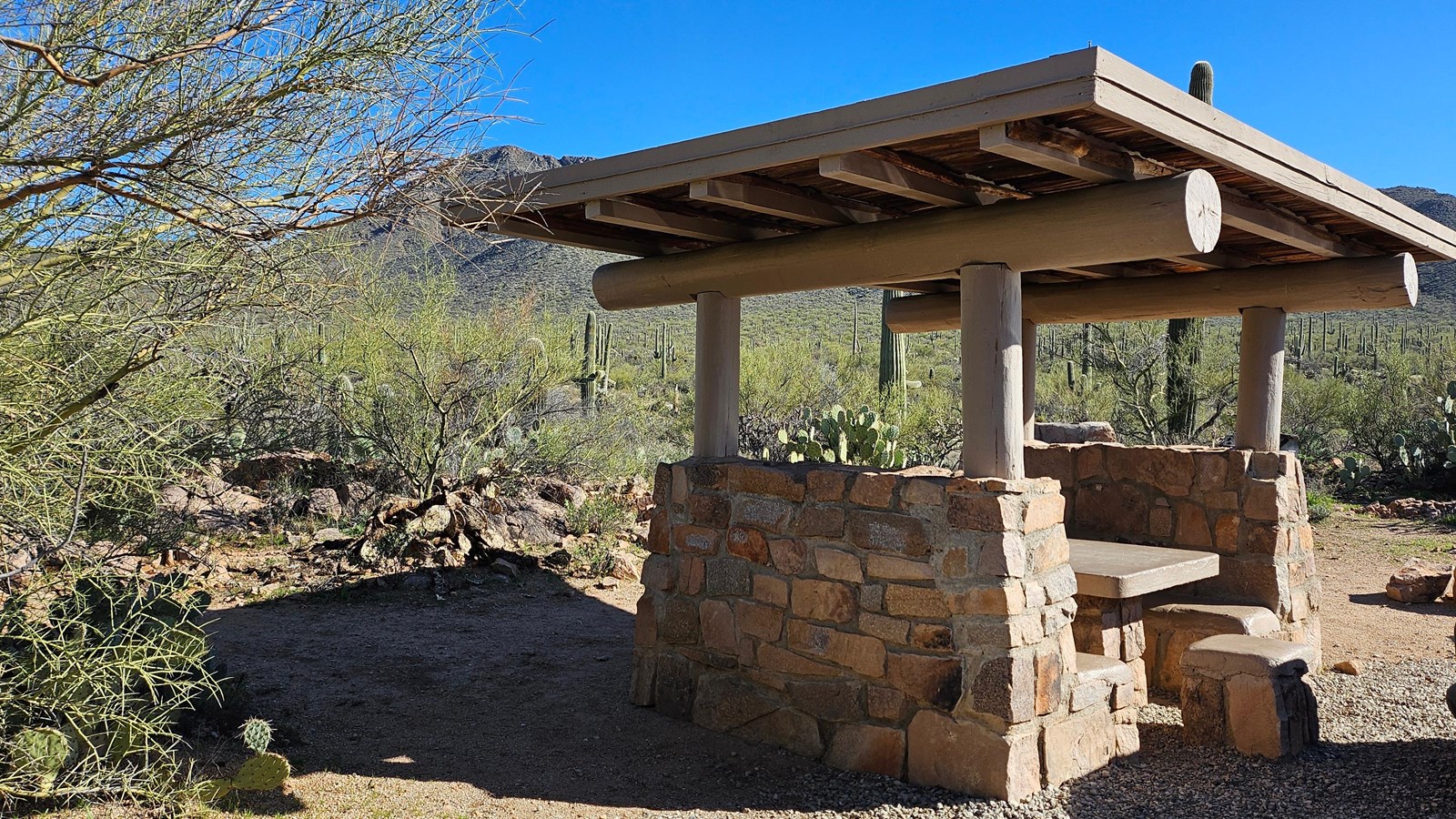 A rock and wood shade structure covers a stone picnic table, surrounded by a lush desert landscape.