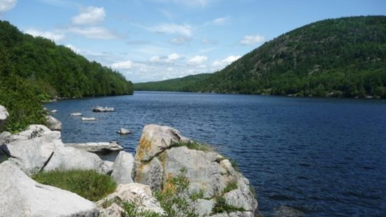 Large rocks in the foreground of a lake bordered by tree-covered hills