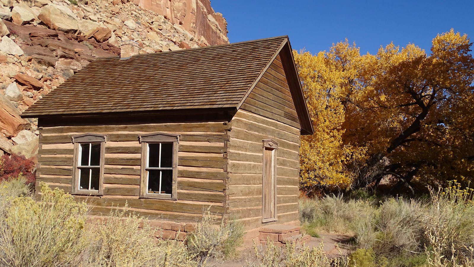 Small log cabin with peaked roof, two windows, and a door, in front of blue sky and red cliffs.