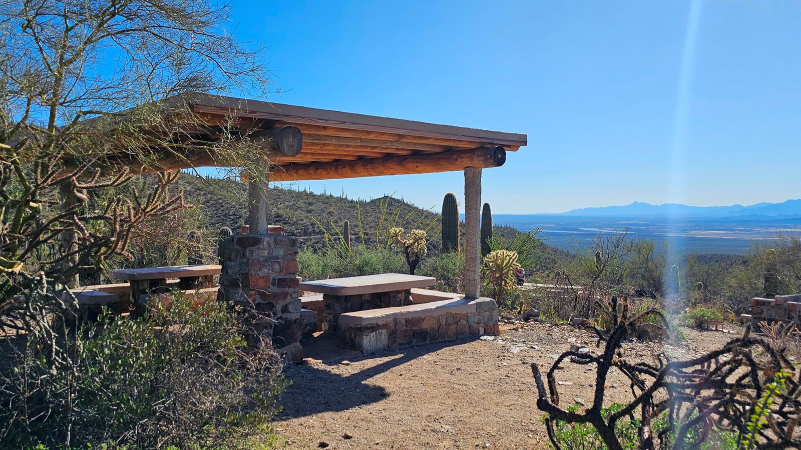 A large wood and stone shade structure covers two stone tables (left). To the right spans a valley.