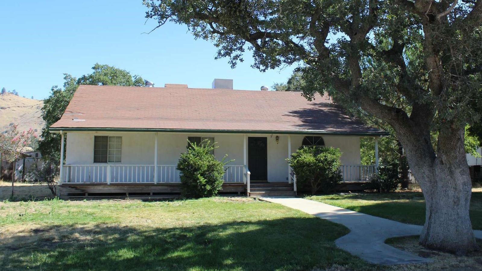 A white house with a lawn, covered porch, and large shade tree.