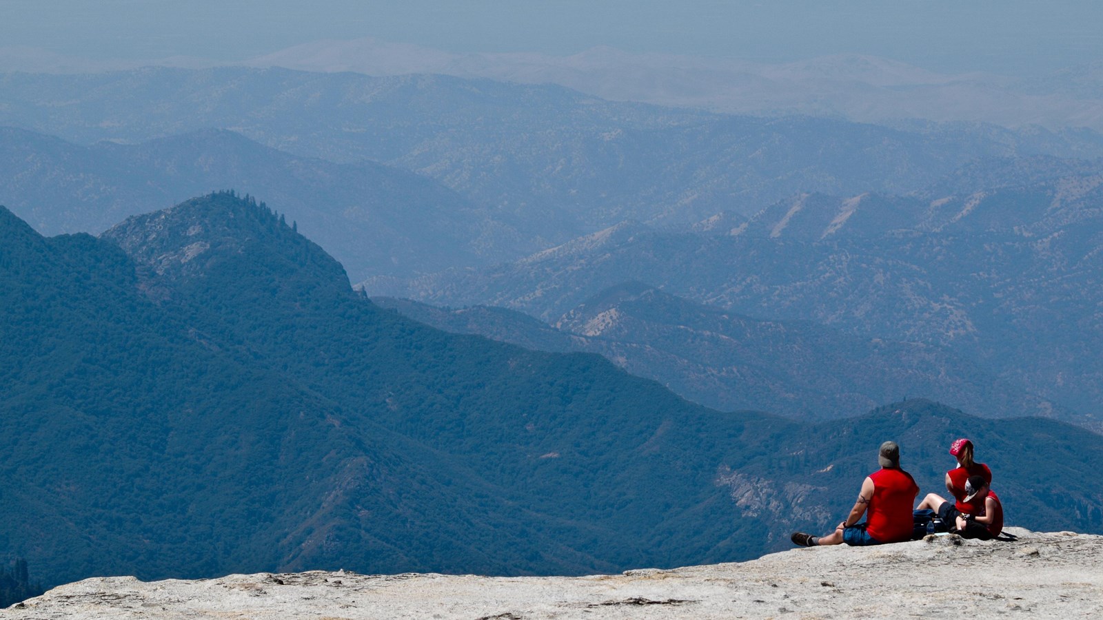 Two people in red sit on the summit of Little Baldy with mountains in the distance.