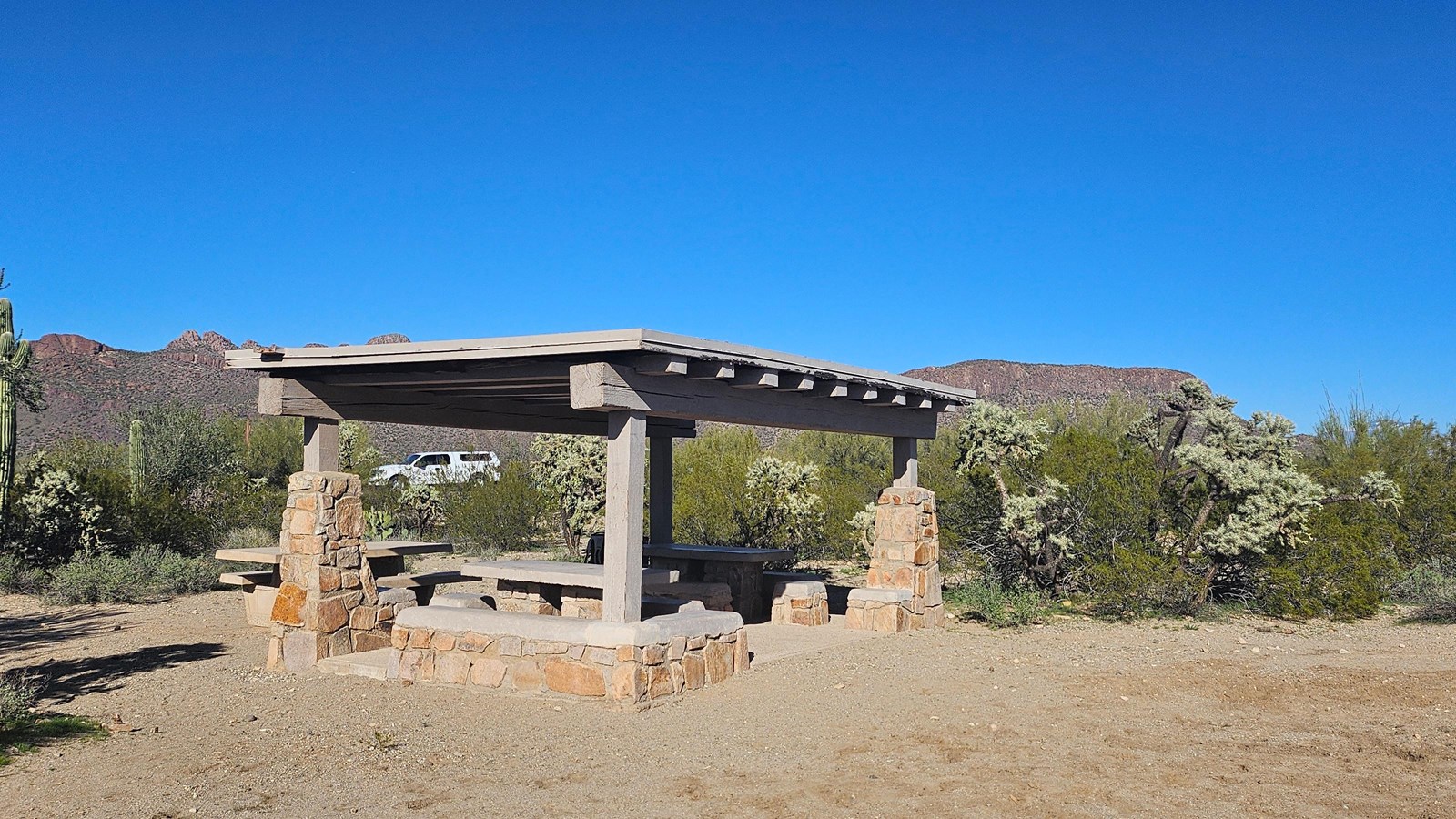 A large stone and wood structure shades two stone tables, with another stone table is uncovered.