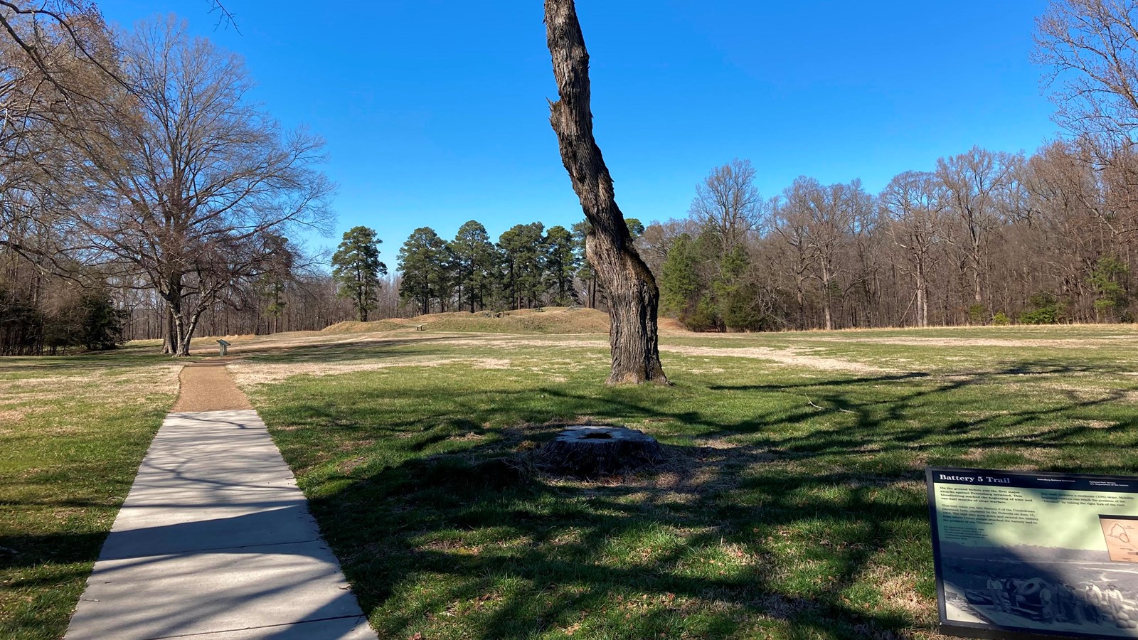 A sidewalk next to a low sign, leads past a tree toward earthworks in the distance. 