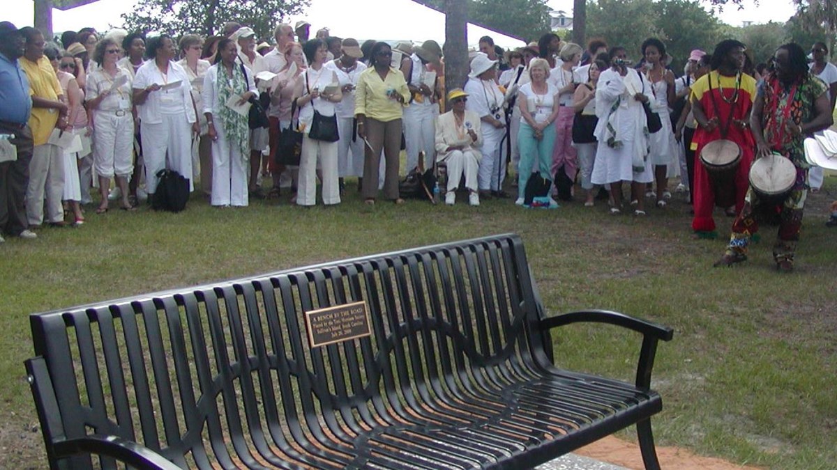 A black metal bench with a plaque is in the foreground with a crowd of people behind it.