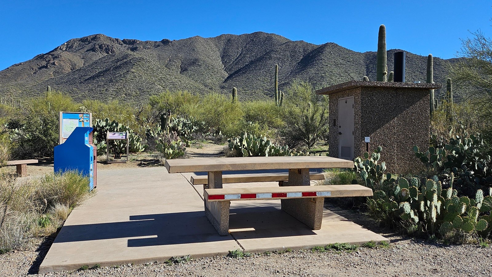A single unshaded, wheelchair accessible, stone table sits on a slap of concrete.