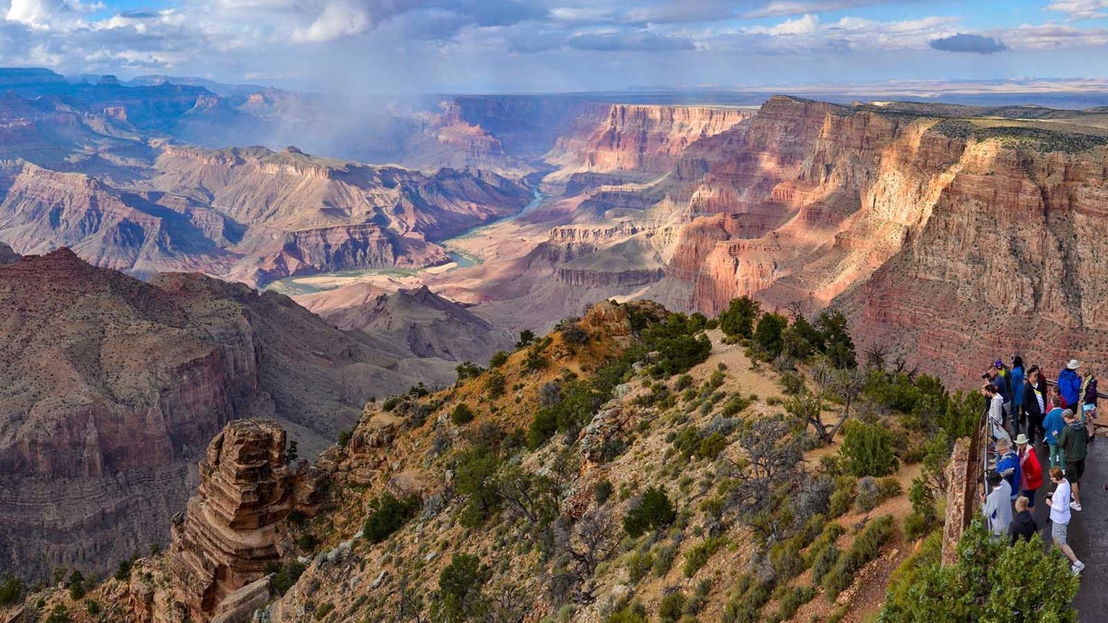 A group of sightseers behind a guardrail at a scenic overlook above a vast and colorful canyon.