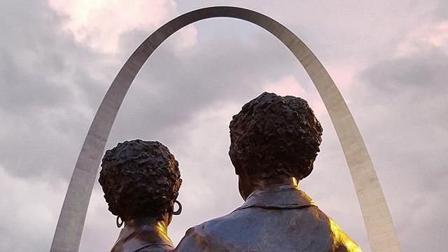 the back of the statue of a man and woman embracing and the Gateway Arch in the background
