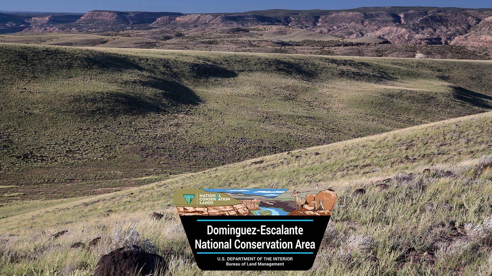 Rolling hills lead to distant desert canyons. Logo sign in foreground.
