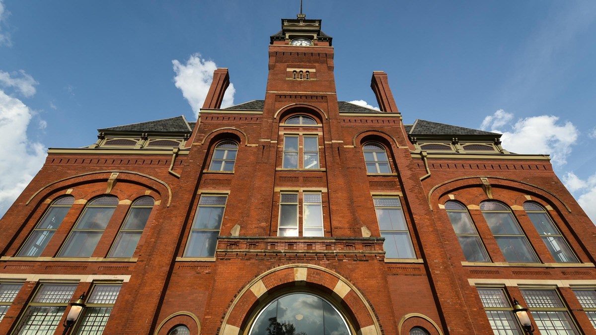 A red brick four story building with a clock tower stands tall against a partly cloudy sky.
