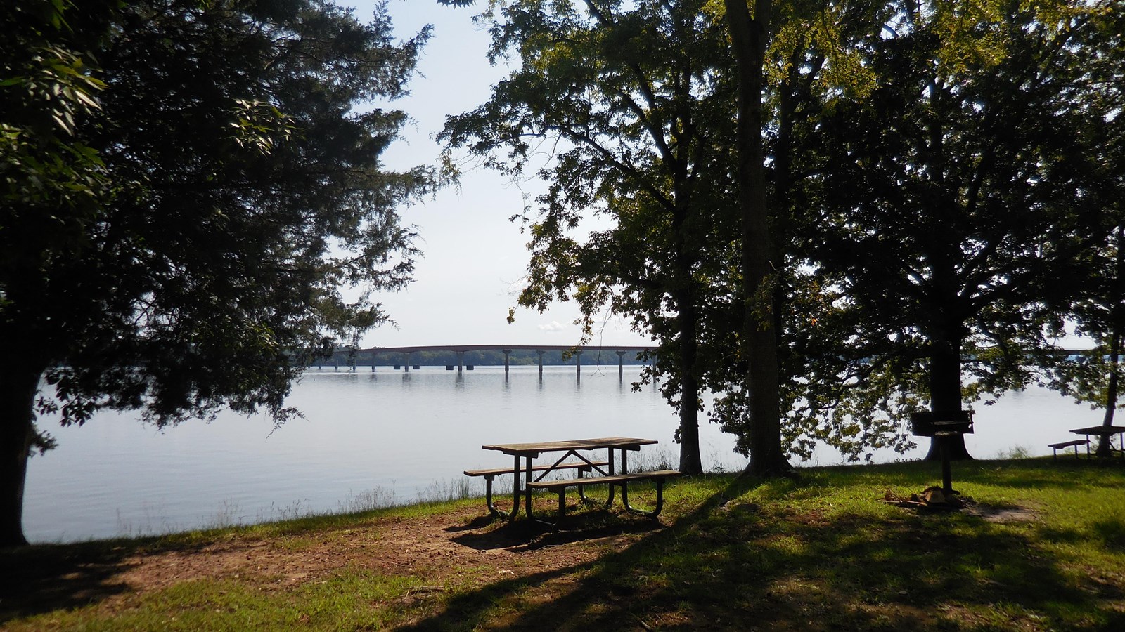 A shaded picnic table in front of a wide river with a long bridge.