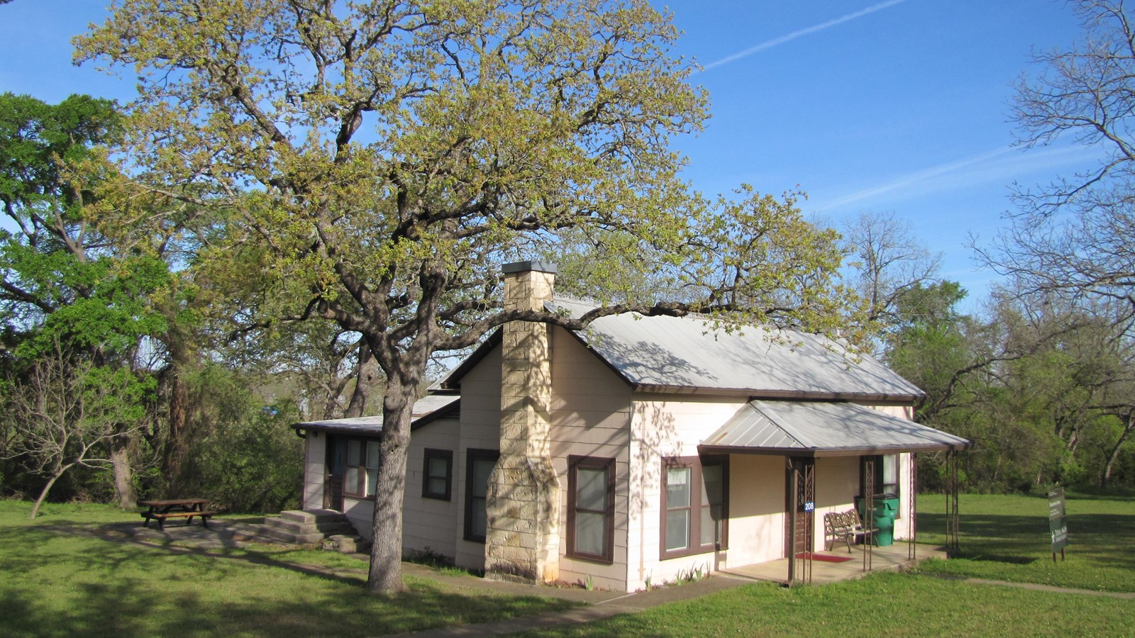 A modest home with a front porch and chimney sits in a small clearing surrounded by trees.