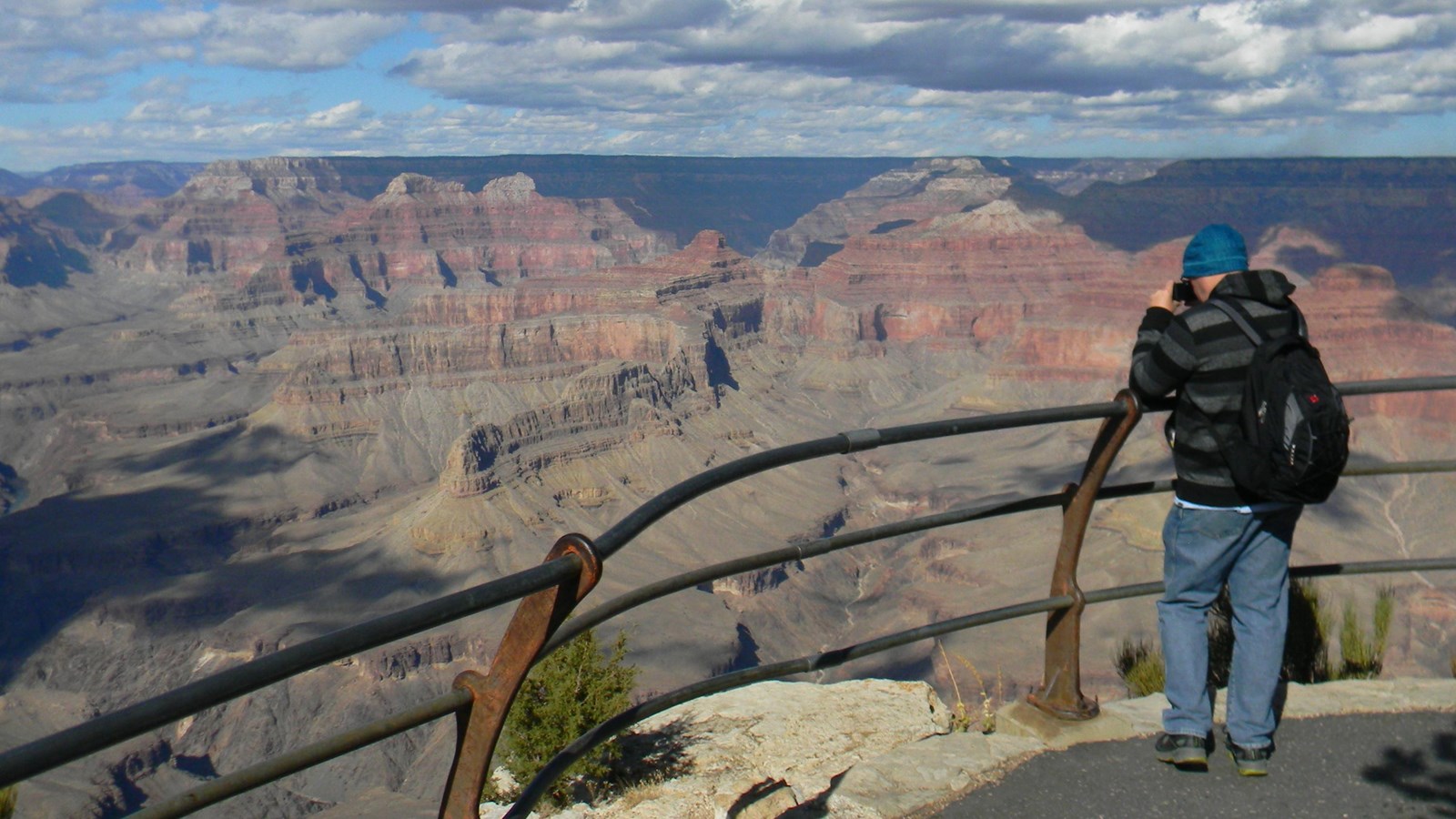 A person leans across a railing and takes a photo of the canyon landscape beyond