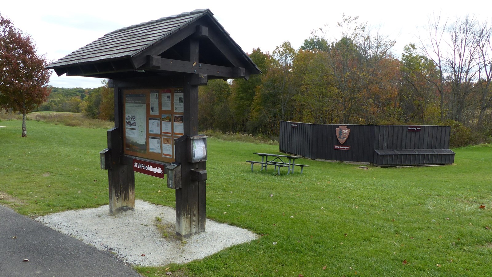 Brown bulletin board with roof and map boxes by a paved trail. Brown fence and picnic table behind.