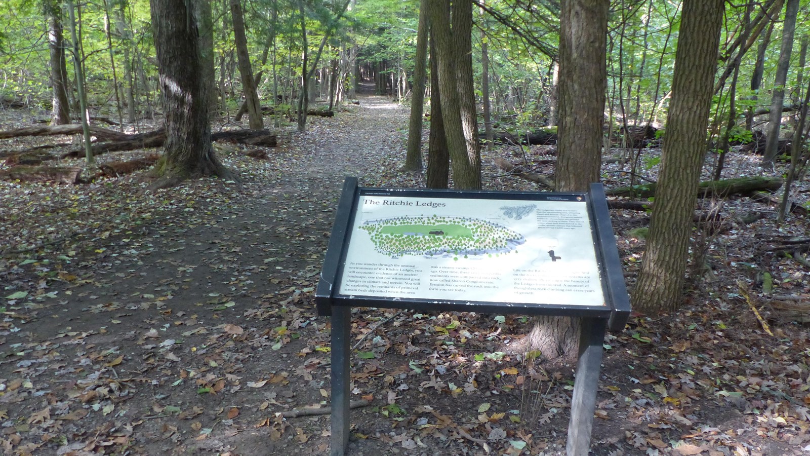 “Ritchie Ledges” panel stands along an unpaved trail surrounded by tree trunks and fallen leaves.