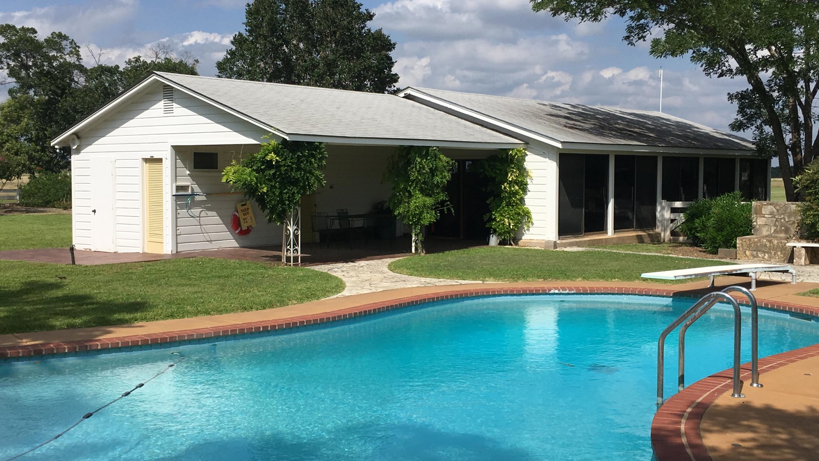 Behind a blue swimming pool, there\'s a small white building with yellow doors and dark windows.