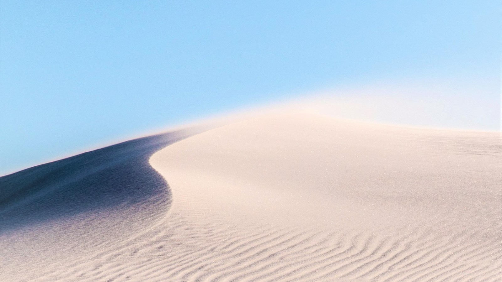 Rippled white sand dune with wind blowing over the crest of the dune. Half the dune is in shadow.