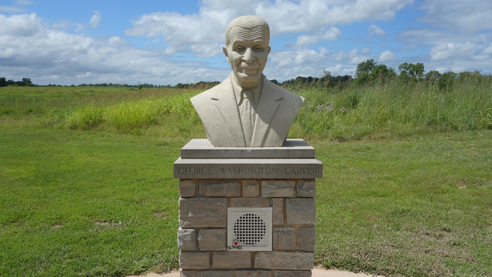 Cast concrete bust of George Washington Carver on a cut stone cap over a concrete block base.