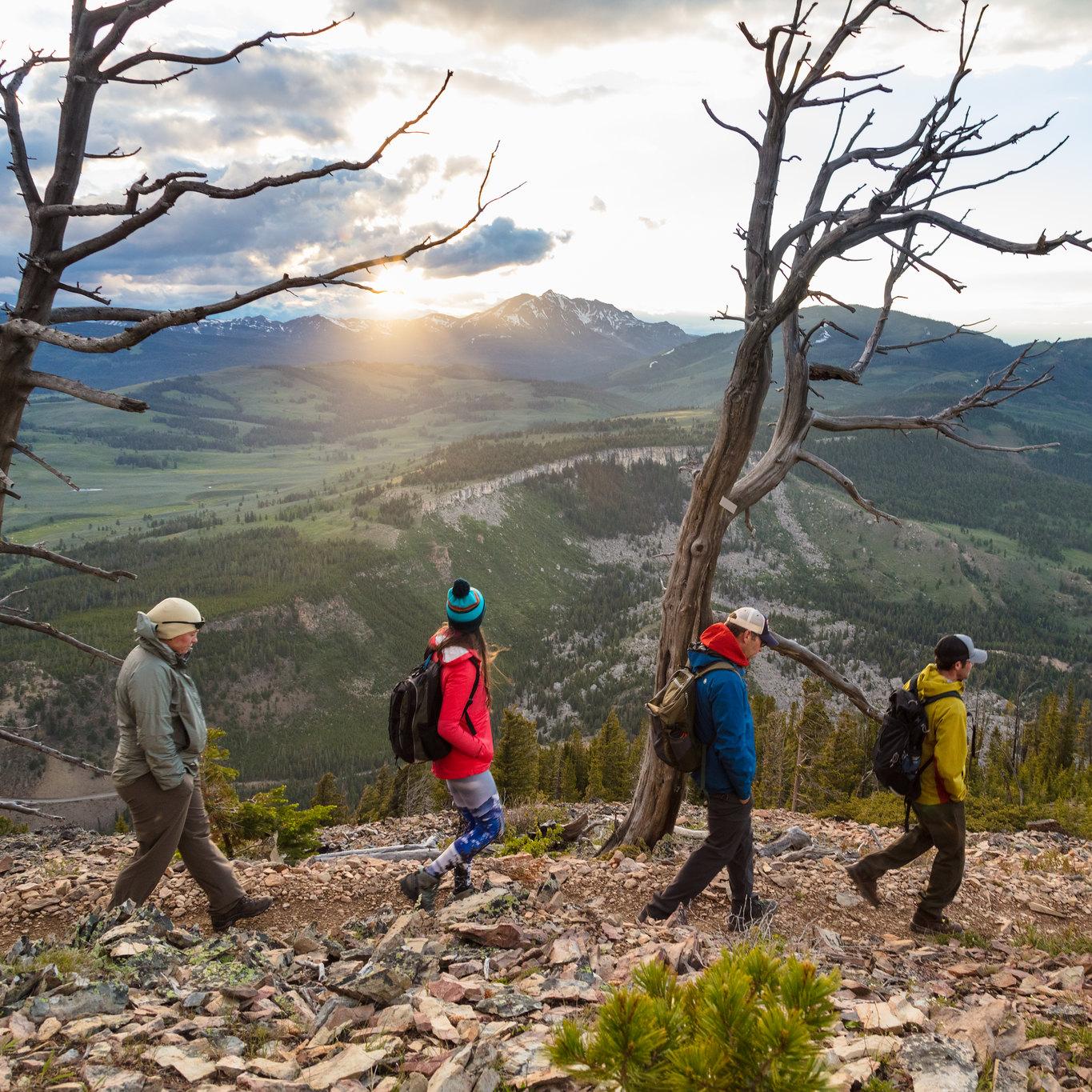 Four people hike across a talus slope with the sun setting behind a mountain in the distance.